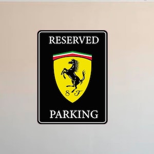 Puede incluir: Se&ntilde;al rectangular negra de "RESERVED PARKING". La se&ntilde;al presenta el logotipo de Ferrari: un caballo negro encabritado sobre un escudo amarillo con las letras "SF" y una franja verde, blanca y roja. Montada en una pared beige.