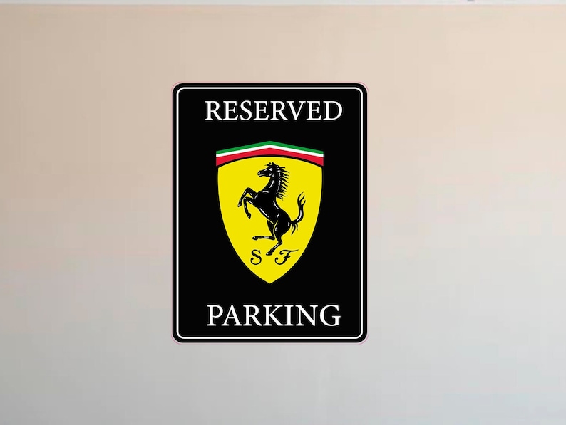 Puede incluir: Se&ntilde;al rectangular negra de "RESERVED PARKING". La se&ntilde;al presenta el logotipo de Ferrari: un caballo negro encabritado sobre un escudo amarillo con las letras "SF" y una franja verde, blanca y roja. Montada en una pared beige.