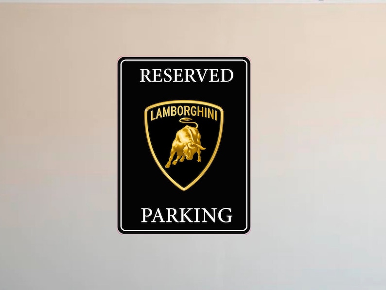 Puede incluir: Cartel rectangular negro con texto blanco &laquo;&nbsp;RESERVED PARKING&nbsp;&raquo;. El centro presenta el logotipo de Lamborghini en oro, un escudo con un toro dorado. El cartel tiene un borde negro.
