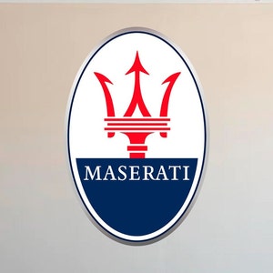Puede incluir: Logotipo ovalado de Maserati con borde plateado. El logotipo presenta un tridente rojo sobre un fondo blanco, con la palabra "MASERATI" en azul marino en la parte inferior.