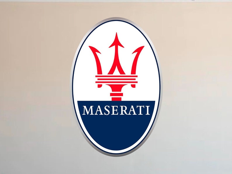 Puede incluir: Logotipo ovalado de Maserati con borde plateado. El logotipo presenta un tridente rojo sobre un fondo blanco, con la palabra "MASERATI" en azul marino en la parte inferior.