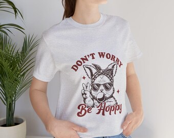 以下が含まれることがあります： バンダナとサングラスをかけた漫画のウサギが描かれたグレーのTシャツ。ウサギはピースサインを出しており、Tシャツには「Don't Worry Be Hoppy」という文字がプリントされています。