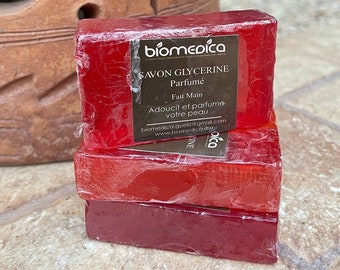 May include: Three bars of red glycerin soap with the text "BIOMEDICA SAVON GLYCERINE Parfumé Fait Main Adoucit et parfume votre peau biomedica.gueliz@gmail.com www.biomedica.ma" printed on the top bar.