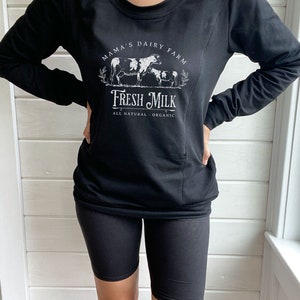May include: Black long-sleeve sweatshirt with a white graphic design that says "Mama's Dairy Farm Fresh Milk All Natural - Organic" with a drawing of cows. The person is wearing black biker shorts.
