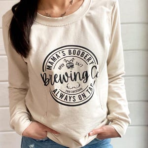 Beige Crewneck Sweatshirt with a logo in black Mama Boobery Brewing Co. Always on Tap 24/7 Two Locations. Breastfeeding Friendly Sweatshirt with zippers on either side that zip from the bottom up for easy accessibility to latch baby to nurse.
