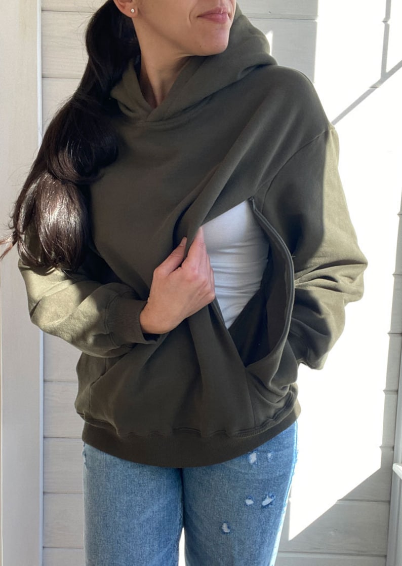 Pu&ograve; includere: Una felpa con cappuccio verde scuro con una cerniera nascosta per un facile allattamento al seno. La felpa &egrave; mostrata indossata con una canottiera bianca e jeans blu.