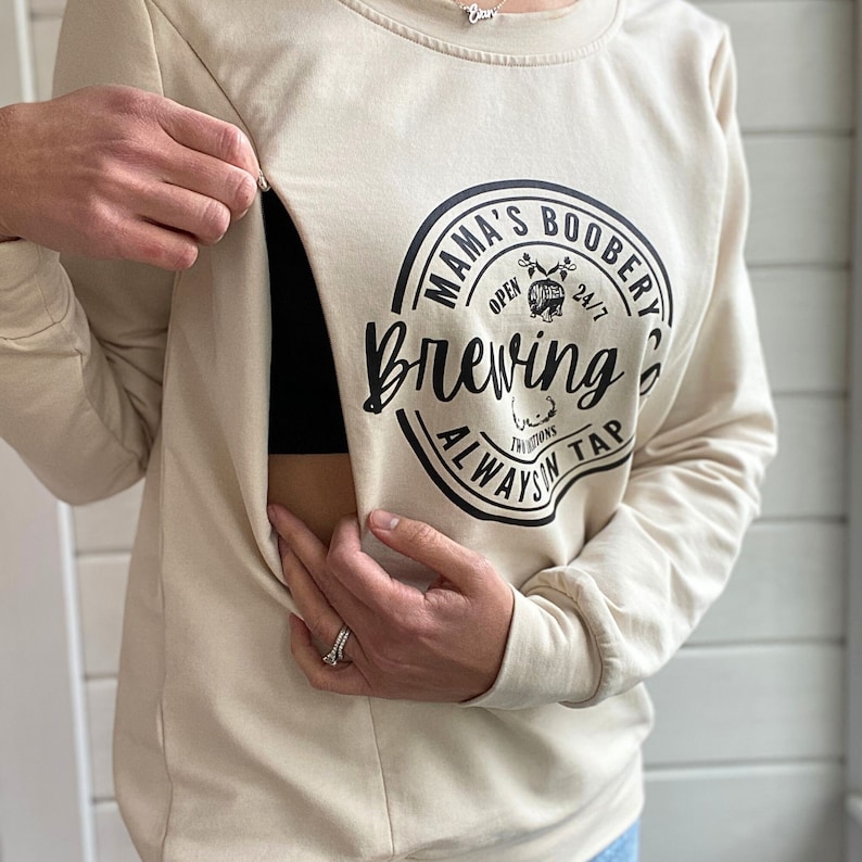 Beige Crewneck Sweatshirt with a logo in black Mama Boobery Brewing Co. Always on Tap 24/7 Two Locations. Breastfeeding Friendly Sweatshirt with zippers on either side that zip from the bottom up for easy accessibility to latch baby to nurse.