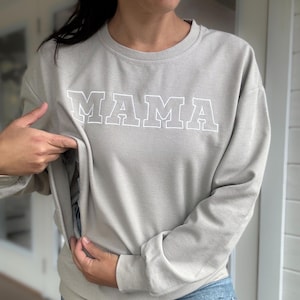 May include: A light gray sweatshirt with the word "MAMA" in white block letters. The sweatshirt has a ribbed texture and a relaxed fit. The design is simple and modern, suitable for casual wear.