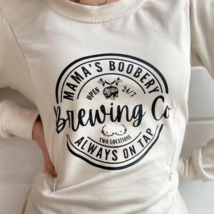 Beige Crewneck Sweatshirt with a logo in black Mama Boobery Brewing Co. Always on Tap 24/7 Two Locations. Breastfeeding Friendly Sweatshirt with zippers on either side that zip from the bottom up for easy accessibility to latch baby to nurse.