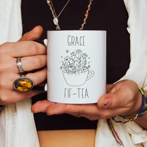 May include: White ceramic mug with black interior, featuring the text "GRACE FIF-TEA" and a floral illustration inside a teacup. The mug is held by a person wearing rings and bracelets.