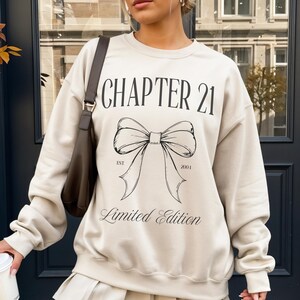 May include: Sand-colored sweatshirt with the text "CHAPTER 21" above a bow graphic, with "EST 2001" and "Limited Edition" below. The sweatshirt is paired with a pleated skirt and white boots.