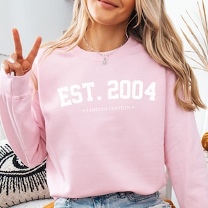 May include: A light pink sweatshirt with the text "EST. 2004" in white, and the words "LIMITED EDITION" in smaller white text below.