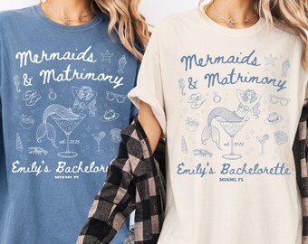May include: Two t-shirts with a blue mermaid design and the text "Mermaids & Matrimony" in white. The text "Emily's Bachelorette" is below the design on the ivory t-shirt and "Emily's Bachelor" is below the design on the blue jean t-shirt.