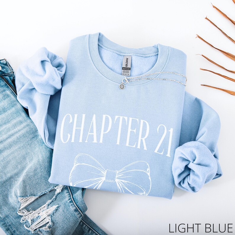 May include: A light blue sweatshirt with the text "CHAPTER 21" and a bow graphic. A silver necklace is draped across the sweatshirt, which is laid on top of a pair of blue jeans. The words "LIGHT BLUE" are at the bottom.