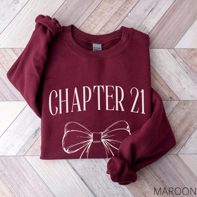 May include: Maroon sweatshirt with the text "CHAPTER 21" in white, along with a white bow graphic. The sweatshirt is made of a soft material and is displayed on a wooden surface.