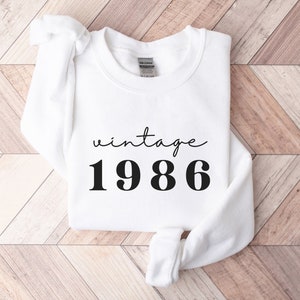 May include: White sweatshirt with black text that reads "vintage 1986".