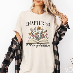 May include: A white t-shirt with the text "Chapter 30 A Literary Milestone" and a floral illustration of a book with flowers.