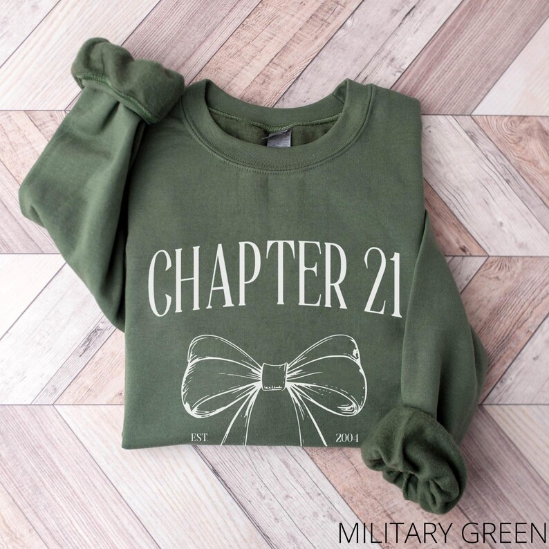 May include: A military green sweatshirt with the text "CHAPTER 21" in white, along with a bow graphic and the year "2004". The sweatshirt is folded, with the sleeves visible. The background is a light brown wooden surface.