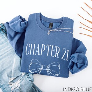 May include: Indigo blue sweatshirt with the text "CHAPTER 21" and a bow graphic. A silver necklace with a pendant is draped on the sweatshirt. The sweatshirt is paired with blue jeans.