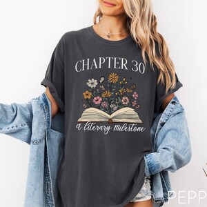 May include: A dark gray t-shirt with the text "Chapter 30 a literary milestone" and an illustration of an open book with flowers growing out of it.