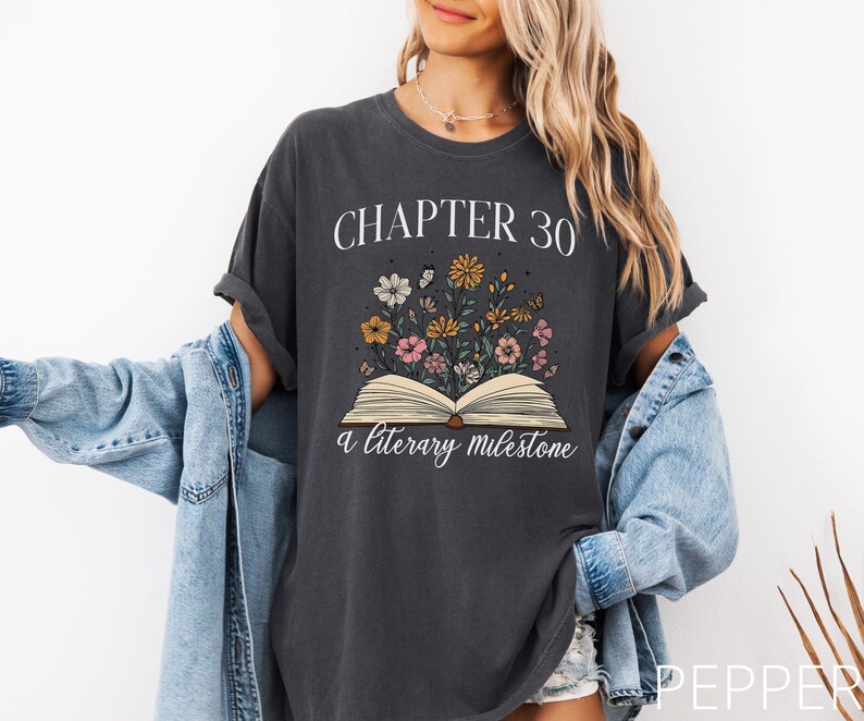 May include: A dark gray t-shirt with the text "Chapter 30 a literary milestone" and an illustration of an open book with flowers growing out of it.