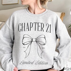 May include: A light gray sweatshirt with the text "CHAPTER 21" above a large bow graphic. Below the bow, the text reads "EST 2001" and "Limited Edition". The sweatshirt is a casual style.