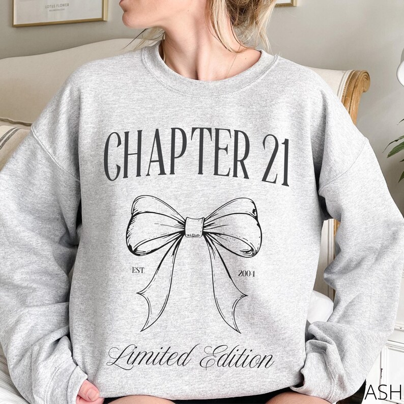 May include: A light gray sweatshirt with the text "CHAPTER 21" above a large bow graphic. Below the bow, the text reads "EST 2001" and "Limited Edition". The sweatshirt is a casual style.