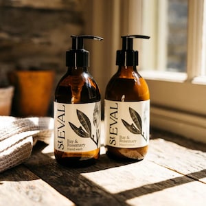 May include: Two brown glass bottles with black pump dispensers. Each bottle has a white label with the text "St Eval Bay & Rosemary" and a botanical illustration. The bottles are on a wooden surface.