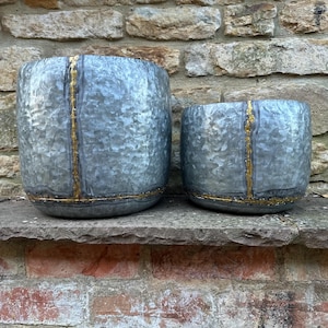 May include: Two galvanised metal planters, one large and one small, sit side-by-side. The planters have a cylindrical shape with a textured surface and gold-coloured accents. Suitable for indoor or outdoor use.