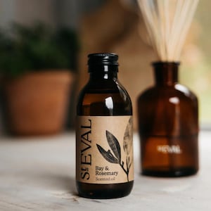 May include: A brown glass bottle of St Eval Bay & Rosemary scented oil with a black cap. The label features a leaf illustration. A brown glass diffuser bottle with reeds is in the background.