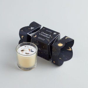 May include: A small glass candle in a clear jar with a black and gold label that says "St Eval Orange & Cinnamon" and a star pattern. The candle is in a festive cracker-style box with a black and gold design.
