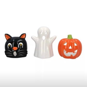 May include: Three ceramic Halloween candle holders. A black cat, a white ghost, and an orange pumpkin with a jack-o'-lantern face.