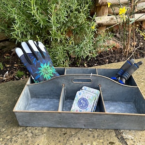 May include: A galvanized metal gardening caddy with four compartments. The caddy contains a pair of gardening gloves with a black and green floral print, a set of seed packets, and a pair of gardening shears.