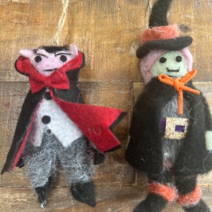 May include: Two felt Halloween ornaments: a vampire with a red bow tie and cape, and a witch with a black hat and orange accents. Both are suspended from twine strings against a wooden backdrop.