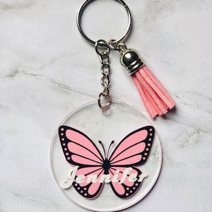 Puede incluir: Un llavero de acrílico transparente con una mariposa rosa y el nombre "Jennifer" en letras blancas. El llavero tiene un anillo plateado y un borla rosa.