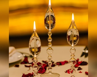 May include: Three clear glass oil lamps with flames burning. The lamps have a teardrop-shaped glass bowl and a decorative beaded stem. The lamps are sitting on a white table with red rose petals scattered around them.