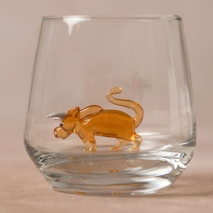 May include: A small, amber-colored glass figurine of a triceratops dinosaur inside a clear glass tumbler.