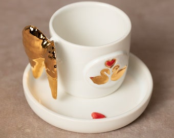 May include: White ceramic coffee cup with a gold wing handle and a gold swan design with a red heart. The cup is on a matching saucer.