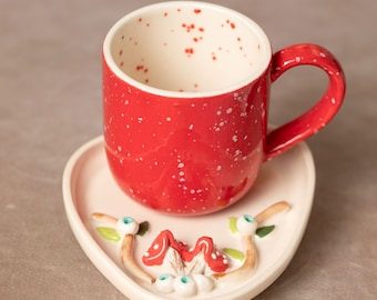 May include: A red ceramic coffee cup with white speckles and a white ceramic saucer with a mushroom design. The saucer is heart-shaped.