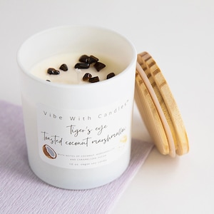 May include: White candle jar with a wooden lid. The candle is labeled "Vibe With Candles" and "Tiger's eye Toasted coconut marshmallow" with notes of coconut, marshmallow, and caramelized sugar. The candle is 10 oz and vegan soy.