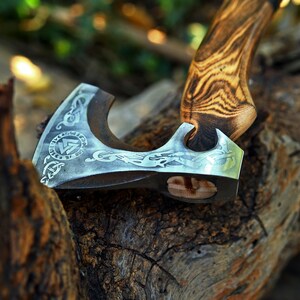 May include: A hand-forged axe with a wooden handle and a steel head. The head is decorated with intricate carvings and a symbol. The axe is resting on a piece of weathered wood.