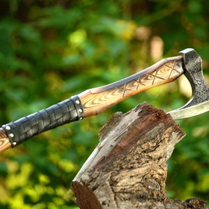 May include: A hand-forged axe with a wooden handle wrapped in black leather. The axe head has a hammered finish and a sharp blade. The handle is decorated with a carved pattern.