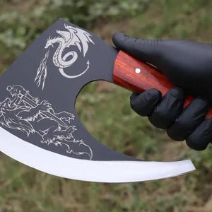 Könnte beinhalten: Axt mit einer dunkelgrauen Klinge und einem polierten Holzgriff. Die Klinge zeigt ein weißes Drachen- und Wolfsdesign. Der Griff ist rotbraun und wird von einer behandschuhten Hand gehalten.