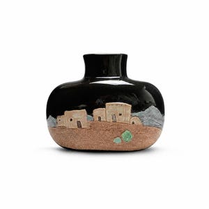 Può includere: Un vaso in ceramica di forma ovale e tozza con una sezione superiore nera lucida e una sezione inferiore color marrone chiaro opaco e testurizzato. La parte anteriore presenta un rilievo di edifici in adobe, cime montuose blu e due piccoli accenti circolari verdi.