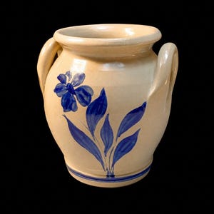 May include: A cream-colored ceramic crock featuring two side handles and a hand-painted blue floral design on the front. The vessel has a wide, rounded rim and a circular base with a thin blue horizontal line encircling the bottom section.