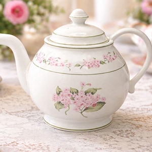 Könnte beinhalten: Weiße Keramik-Teekanne mit Deckel und Henkel, verziert mit rosa Blumenmustern und grünen Akzenten. Die Teekanne hat einen abgerundeten Körper und einen Ausguss. Eine passende Tasse und Untertasse befinden sich im Hintergrund.