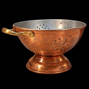 May include: A round copper colander featuring a hammered exterior finish, a silver-toned interior, and two polished brass side handles. The bowl is supported by a circular pedestal base and is perforated with numerous small drainage holes.