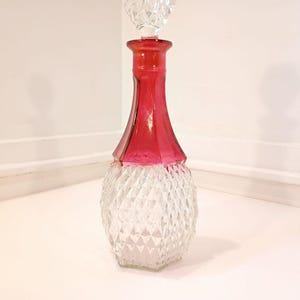 May include: A vintage glass decanter with a ruby red neck and a clear, diamond-cut base. The decanter has a crystal stopper. This decorative piece is suitable for serving wine or spirits.