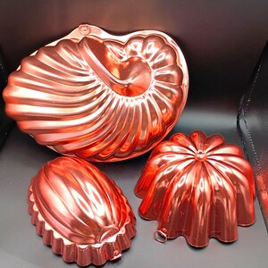 May include: Three copper-colored vintage jello molds in various shapes. The largest mold is a nautilus shell design. The other two molds are a pumpkin and a teardrop shape. All molds have a shiny, reflective surface.