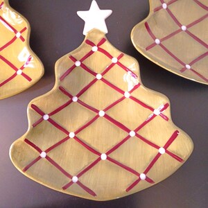May include: Christmas tree-shaped serving plates. The plates are a golden yellow color with a red crisscross pattern and white dots. Each plate has a white star at the top. Festive holiday decor.